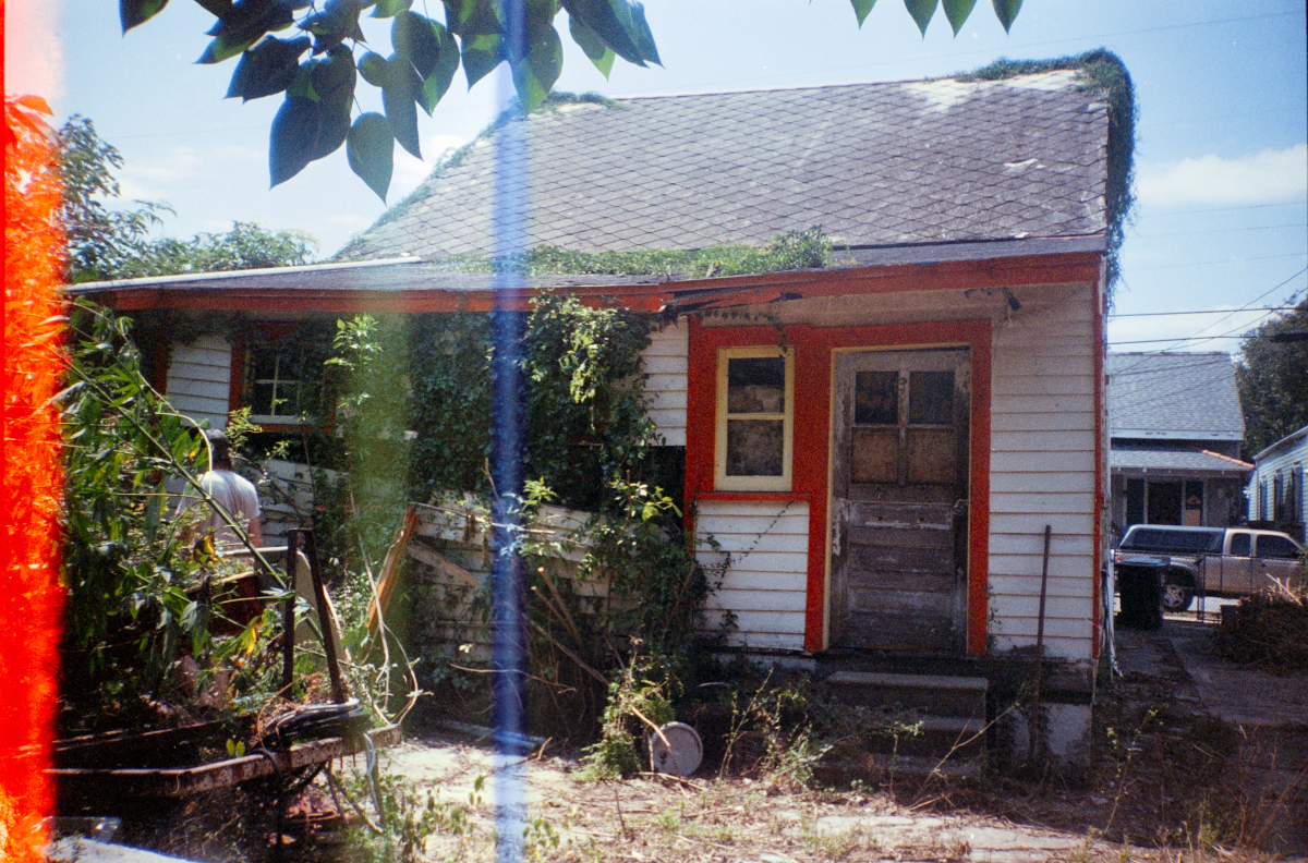 film photo of an overgrown, aging house with a weathered exterior. Vines and plants cover parts of the structure. A figure stands near the house, surrounded by dense bushes. The film has overexposed stripes and lots of grain- imperfect beautiful photo
