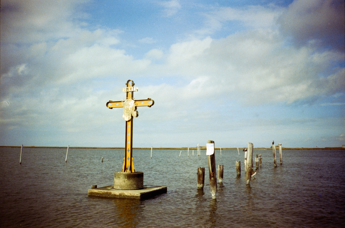 a grainy film photo of a large cross in shallow water, with surrounding wooden posts. The sky is partly cloudy, the atmosphere is serene.